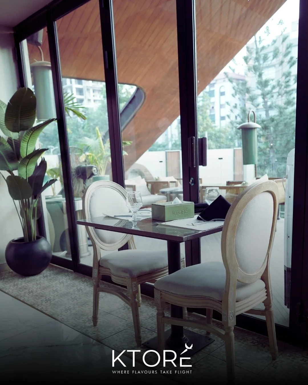 A table for two by floor-to-ceiling glass, cane-backed chairs and palms